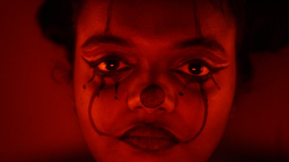 A person with clown makeup featuring exaggerated eyes and a red nose, illuminated by a strong red light. The makeup includes black tears and a painted smile contrasted against dark shadows.