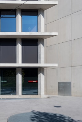 A modern building facade with large, square windows arranged in a grid pattern. The structure is made of concrete with a smooth finish and features a minimalist design. The glass windows reflect a clear blue sky while some are covered with dark blinds. A red sign labeled 'UniFor' is displayed on the window near the entrance. The ground is paved with concrete tiles, and a round metal grate is visible in the foreground.