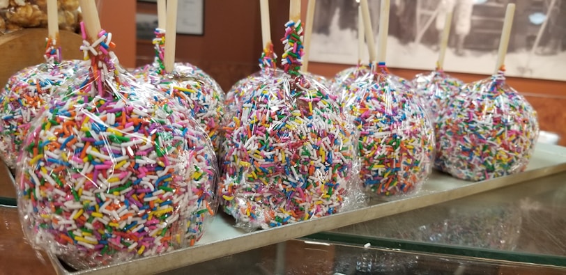 Several candy apples covered in colorful sprinkles are arranged in a line on a tray. Each apple is wrapped in clear plastic and has a wooden stick for holding.