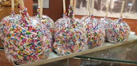 Several candy apples covered in colorful sprinkles are arranged in a line on a tray. Each apple is wrapped in clear plastic and has a wooden stick for holding.