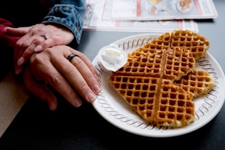 A plate of heart-shaped waffles is served with a small container of butter, placed on a patterned plate. Beside the plate, two hands are gently clasped, possibly conveying a sense of warmth and connection. The setting includes a table with partially visible menus.