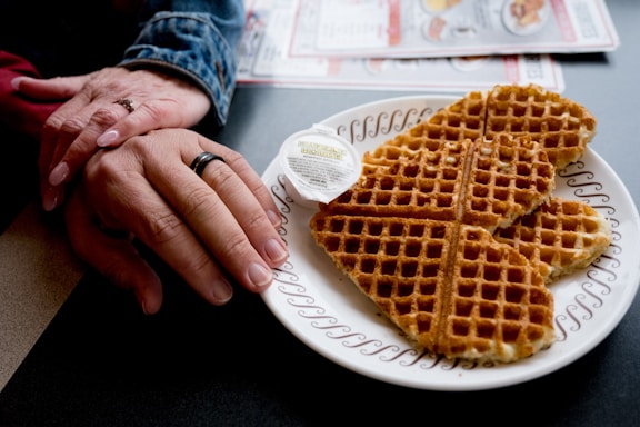 A plate of heart-shaped waffles is served with a small container of butter, placed on a patterned plate. Beside the plate, two hands are gently clasped, possibly conveying a sense of warmth and connection. The setting includes a table with partially visible menus.