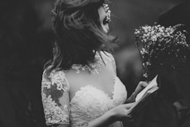 A woman in a lacy dress is holding a bouquet of small flowers and an open book. Her hair is gently blowing across her face, and the scene is captured in black and white, enhancing the romantic and vintage tone.
