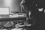 A person deeply focused on working at a music production setup, with a laptop and other electronic equipment on a desk. The lighting is low, creating dramatic shadows and highlighting the concentrated expression on the person's face.