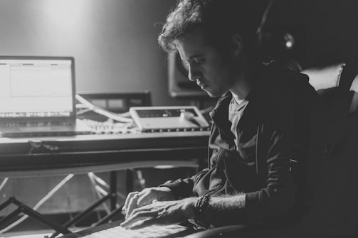 A person deeply focused on working at a music production setup, with a laptop and other electronic equipment on a desk. The lighting is low, creating dramatic shadows and highlighting the concentrated expression on the person's face.