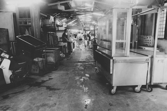 A black and white image of a narrow marketplace aisle lined with various stalls and equipment. Some people are visible in the distance, walking or standing under a canopy, creating a busy yet blurred background. The atmosphere seems cluttered with various items like a motorcycle, carts, and display cases scattered around.