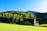 A quaint church with a peaked roof sits in a lush, green meadow surrounded by a dense forest of towering evergreen trees. The background is dominated by a range of gently sloping hills under a clear blue sky, evoking a serene and picturesque rural landscape.