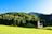 A quaint church with a peaked roof sits in a lush, green meadow surrounded by a dense forest of towering evergreen trees. The background is dominated by a range of gently sloping hills under a clear blue sky, evoking a serene and picturesque rural landscape.