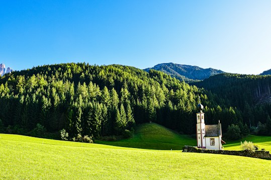 A quaint church with a peaked roof sits in a lush, green meadow surrounded by a dense forest of towering evergreen trees. The background is dominated by a range of gently sloping hills under a clear blue sky, evoking a serene and picturesque rural landscape.