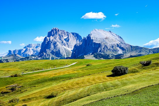A picturesque landscape featuring a vast green meadow with a winding path cutting across it. In the background, towering mountains with rugged, rocky faces rise majestically under a clear blue sky. A few small clouds dot the sky, adding to the serenity of the scene.