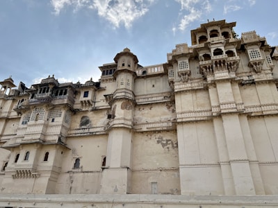 The image depicts a large, historic building with intricate architectural details, including multiple balconies, windows with decorative grille work, and a series of protruding structures. The walls exhibit signs of weathering, and the structure features a blend of ornate design elements typical of historic Indian architecture.