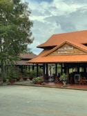 A wooden restaurant building with a red-tiled roof stands surrounded by greenery. The sign indicates the name 'Ben Dinh Restaurant' in both English and another language. Several potted plants are placed around the entrance, and wooden chairs can be seen inside the open-air dining area. Overhead, the sky is partly cloudy.