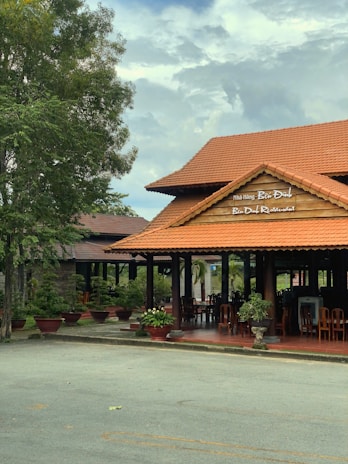 A wooden restaurant building with a red-tiled roof stands surrounded by greenery. The sign indicates the name 'Ben Dinh Restaurant' in both English and another language. Several potted plants are placed around the entrance, and wooden chairs can be seen inside the open-air dining area. Overhead, the sky is partly cloudy.