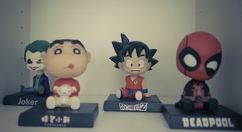 Four figurines are displayed on a shelf. From left to right, there is a Joker figure with green hair and a menacing smile, a round-faced character holding a small animal in a red shirt, a cheerful character with spiky black hair and an orange outfit, and a figure with a mask resembling Deadpool in black and red.