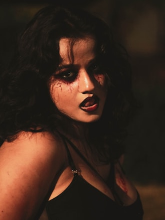 A person with dark hair and intense makeup, featuring dark eyeshadow and lipstick. Red, vein-like details are painted around the eyes, creating a dramatic and eerie appearance. The setting has low lighting, adding a mysterious and gothic vibe.