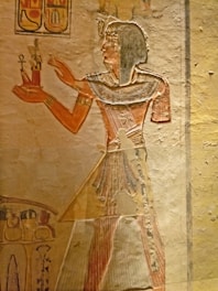 A wall painting showing an ancient Egyptian figure in profile, wearing traditional attire with a headdress. They hold a small object adorned with an ankh and a statue. Hieroglyphics are present above the figure, and additional artwork is visible at the bottom of the frame.