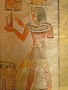 A wall painting showing an ancient Egyptian figure in profile, wearing traditional attire with a headdress. They hold a small object adorned with an ankh and a statue. Hieroglyphics are present above the figure, and additional artwork is visible at the bottom of the frame.