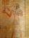 A wall painting showing an ancient Egyptian figure in profile, wearing traditional attire with a headdress. They hold a small object adorned with an ankh and a statue. Hieroglyphics are present above the figure, and additional artwork is visible at the bottom of the frame.