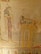 A detailed wall painting depicting ancient Egyptian art. The central figure is a standing human figure, possibly a female, dressed in traditional attire. To the right, there's an assortment of objects, including what appears to be a table with various food items or offerings. The background is made of a textured stone, with hieroglyphs or symbols present in the lower border of the painting.
