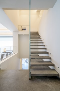 A modern interior staircase with floating slate steps and glass railings. The walls and ceilings are painted in a light cream color, creating a spacious and airy feel. There are visible recessed lights and minimalistic decor, including a chair at the top of the stairs and a table at the landing.