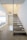 A modern interior staircase with floating slate steps and glass railings. The walls and ceilings are painted in a light cream color, creating a spacious and airy feel. There are visible recessed lights and minimalistic decor, including a chair at the top of the stairs and a table at the landing.