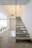 A modern interior staircase with floating slate steps and glass railings. The walls and ceilings are painted in a light cream color, creating a spacious and airy feel. There are visible recessed lights and minimalistic decor, including a chair at the top of the stairs and a table at the landing.