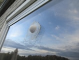 A golf ball is lodged in a cracked window pane, creating an intricate spiderweb pattern of shattered glass around it. The reflection of a partly cloudy sky can be seen in the window, along with some outdoor elements, possibly indicating a backyard or garden setting.
