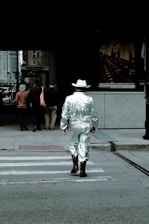 A person wearing a shiny silver suit and a white cowboy hat is walking across a crosswalk in an urban setting. Brown cowboy boots complement the outfit. Other pedestrians are visible ahead on the sidewalk near buildings. The atmosphere suggests a unique blend of modern city life and Western fashion.