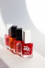 Four nail polish bottles are arranged in a line, each featuring different colors and brands. The bottles have square caps, with the closest one being red and white, followed by an orange, a pink, and another red bottle in the background. The light casts soft shadows behind them, creating a clean and minimalistic setting.