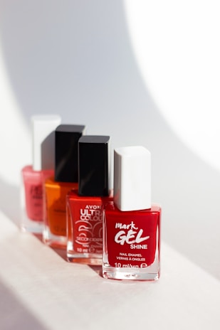 Four nail polish bottles are arranged in a line, each featuring different colors and brands. The bottles have square caps, with the closest one being red and white, followed by an orange, a pink, and another red bottle in the background. The light casts soft shadows behind them, creating a clean and minimalistic setting.
