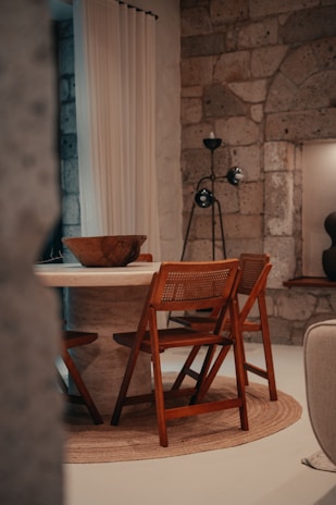 A cozy interior setting featuring a round stone dining table surrounded by wooden chairs with woven seats. The space has a rustic charm with stone walls and a neutral color palette. A tall floor lamp with a unique design stands in the corner, and a large woven rug adds warmth underfoot. Soft natural light filters through sheer curtains, enhancing the serene ambiance.