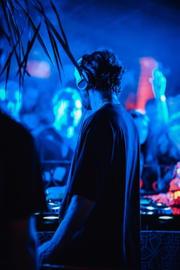 A DJ is performing at a vibrant nightclub filled with blue and red lighting. The atmosphere is lively, with a crowd visible in the background, and the focus is on the DJ wearing headphones, immersed in music.