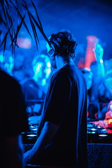 A DJ is performing at a vibrant nightclub filled with blue and red lighting. The atmosphere is lively, with a crowd visible in the background, and the focus is on the DJ wearing headphones, immersed in music.