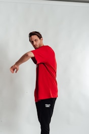 A person wearing a bright red t-shirt and black pants with 'Team USA' written on them poses against a plain, light-colored background. The pose appears dynamic, with the left arm extending outward.
