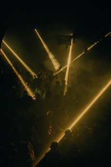Numerous people are gathered in a dark environment illuminated by bright, golden beams of light cutting through the space, creating a dramatic and immersive atmosphere. The scene appears lively with silhouettes of individuals visible amidst the haze, suggesting a party or club event.