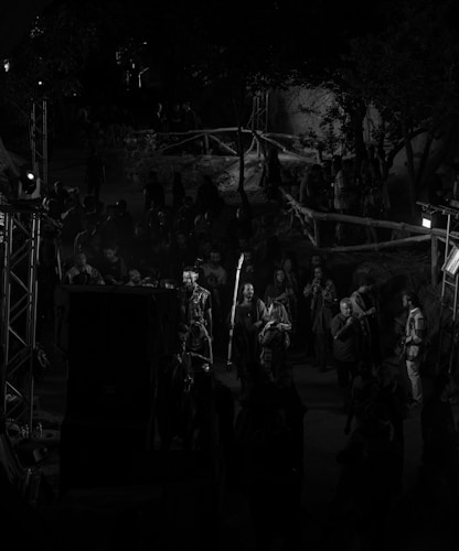 A nighttime event with a crowd of people gathered in an outdoor setting. There are trees and wooden railings in the background, and some individuals are illuminated by spotlights. The atmosphere seems lively with people interacting, some taking pictures and others observing their surroundings.