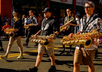 Several people are marching in a parade while playing saxophones. They appear to be part of a marching band and are wearing casual clothing. The sun casts strong shadows on the ground, and a crowd of spectators is visible in the background.
