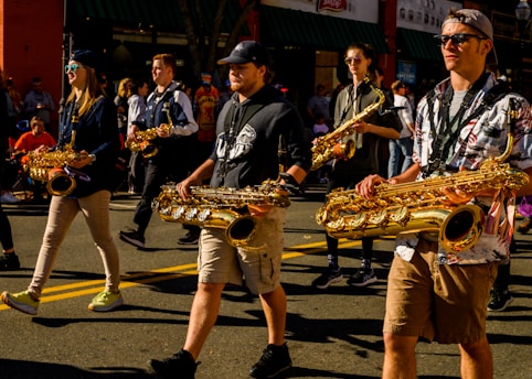 Several people are marching in a parade while playing saxophones. They appear to be part of a marching band and are wearing casual clothing. The sun casts strong shadows on the ground, and a crowd of spectators is visible in the background.