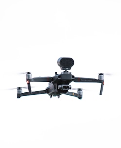 A black and gray quadcopter drone with a camera mounted on the front and extended arms, hovering in a bright sky. The drone has distinct propellers and a modern design.