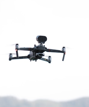 A quadcopter drone hovers in the sky with a camera attached to its underside. It has four rotors and is designed for aerial operations. The background is a clear, light sky.