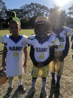 Three young football players in uniforms stand close together outdoors on a grassy field, with one player holding a helmet. The sun shines brightly from the top right corner, casting shadows.