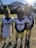 Three young football players in uniforms stand close together outdoors on a grassy field, with one player holding a helmet. The sun shines brightly from the top right corner, casting shadows.
