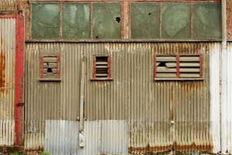 A weathered, corrugated metal wall with patches of rust and peeling paint features three small windows. The window frames are painted red and have broken glass panes, surrounded by worn slats. Above the metal wall is a series of glass panels, some of which are cracked.