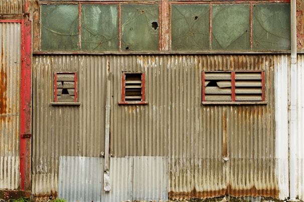 A weathered, corrugated metal wall with patches of rust and peeling paint features three small windows. The window frames are painted red and have broken glass panes, surrounded by worn slats. Above the metal wall is a series of glass panels, some of which are cracked.