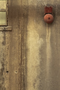 A weathered wall with a rusty, red fire alarm bell. There is a small, frosted window in the top left corner.