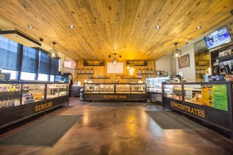 A well-lit store interior featuring wooden ceilings and floors, display cases labeled 'Edibles', 'Flower Bar', and 'Concentrates'. The store has warm lighting with modern hanging lamps and large windows covered with blinds.