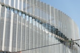 A modern architectural facade featuring a series of vertical metallic panels with a perforated pattern. The panels create a dynamic visual effect through their repetitive geometric arrangement. There is a glass section that reflects the sky and nearby structures, adding depth to the design. In the foreground, a staircase with a railing is visible, and two workers wearing helmets can be seen, suggesting the building is under construction or maintenance.