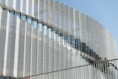 A modern architectural facade featuring a series of vertical metallic panels with a perforated pattern. The panels create a dynamic visual effect through their repetitive geometric arrangement. There is a glass section that reflects the sky and nearby structures, adding depth to the design. In the foreground, a staircase with a railing is visible, and two workers wearing helmets can be seen, suggesting the building is under construction or maintenance.