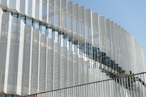 A modern architectural facade featuring a series of vertical metallic panels with a perforated pattern. The panels create a dynamic visual effect through their repetitive geometric arrangement. There is a glass section that reflects the sky and nearby structures, adding depth to the design. In the foreground, a staircase with a railing is visible, and two workers wearing helmets can be seen, suggesting the building is under construction or maintenance.