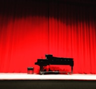 A grand piano and stool are set on a stage with bright red curtains as the backdrop. The lighting highlights the sleek black body of the piano, creating a dramatic and elegant atmosphere.